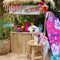 Small white dog in trendy pink Hawaiian shirt at tropical-themed tiki bar with surfboard and fruits, showcasing pet fashion for 2025.