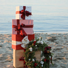 Stack of Christmas gifts wrapped in red and white paper with ribbons, placed on a sandy beach at sunset, decorated with a festive wreath.