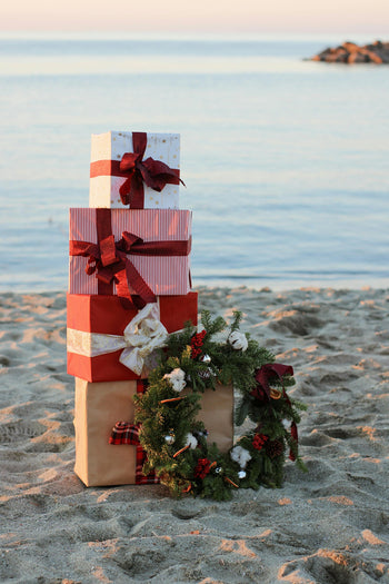 Stack of Christmas gifts wrapped in red and white paper with ribbons, placed on a sandy beach at sunset, decorated with a festive wreath.